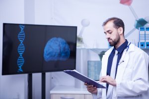 Confident young doctor with a clipboard in his hands standing by a monitor with a brain graph. Handsome doctor.