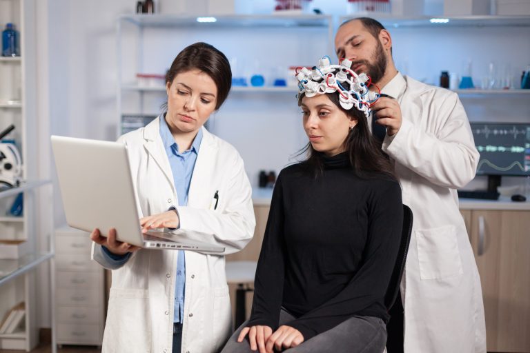 Specialist neurologist doctor taking notes on laptop asking patient's symptoms adjusting high tech eeg headset